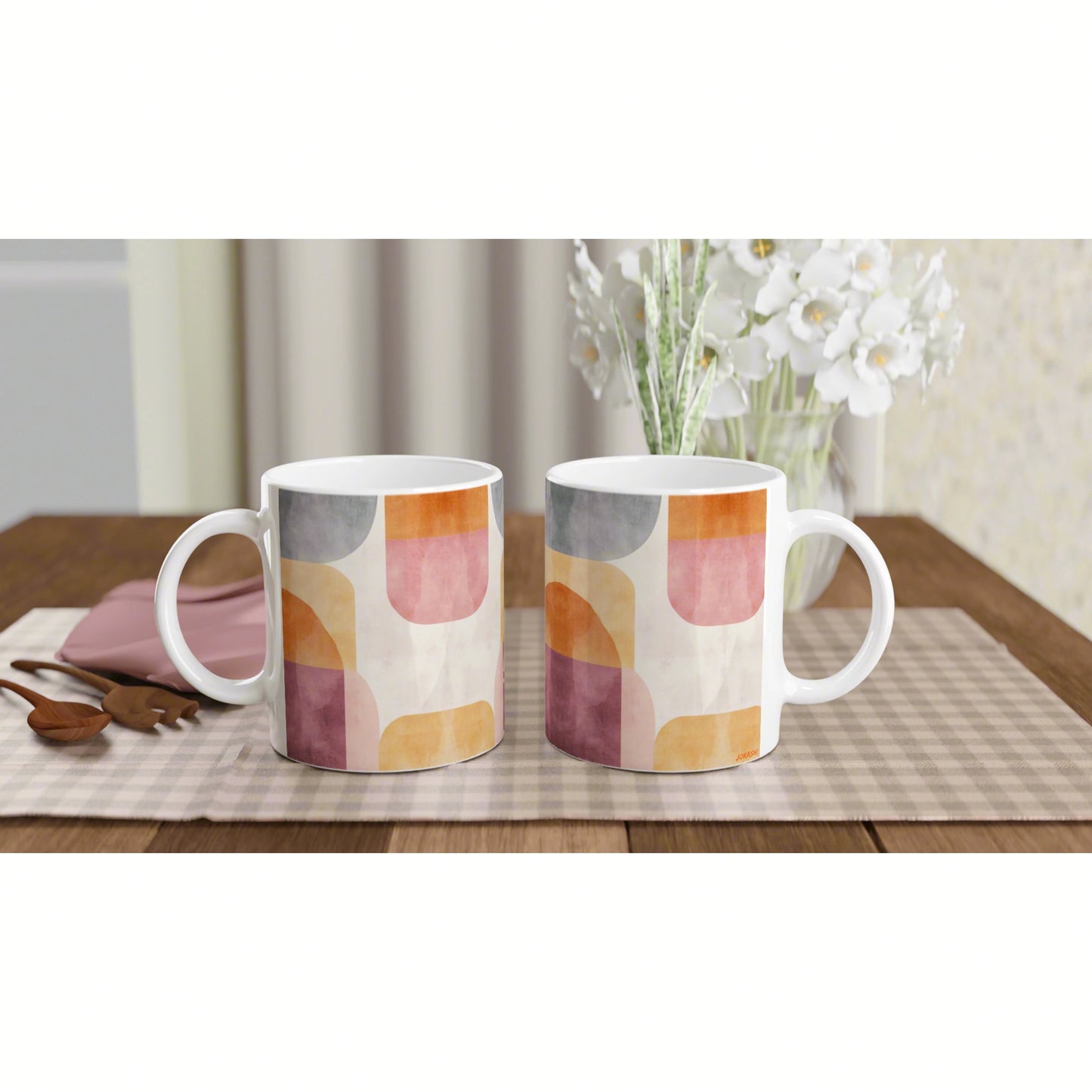 Two white ceramic mugs with a pastel abstract geometric pattern on a wooden table, with white flowers behind.