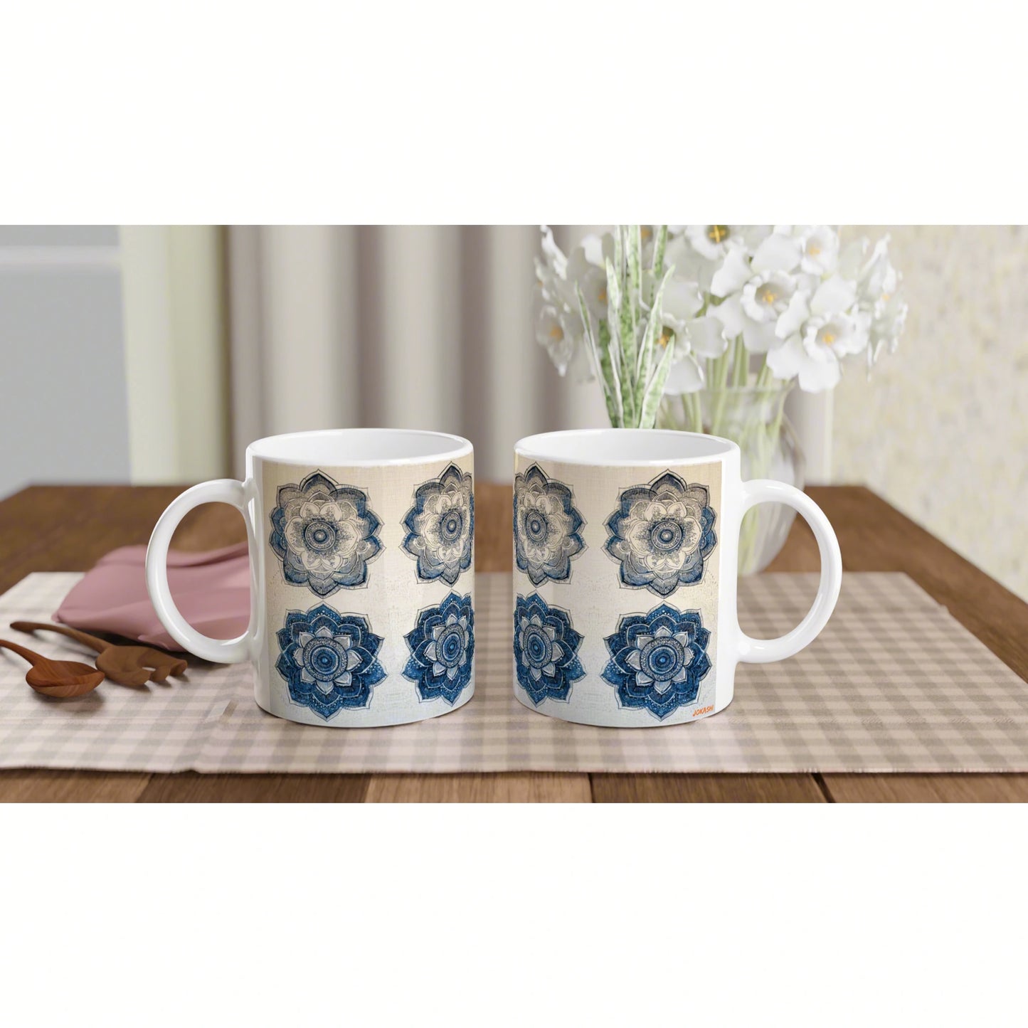 Two white mugs with blue mandala patterns on a beige placemat, on a wooden table with white flowers in the background.