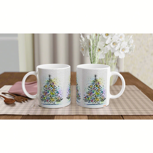 Two white ceramic mugs with colorful watercolor Christmas tree designs on a wooden table with a plaid placemat.
