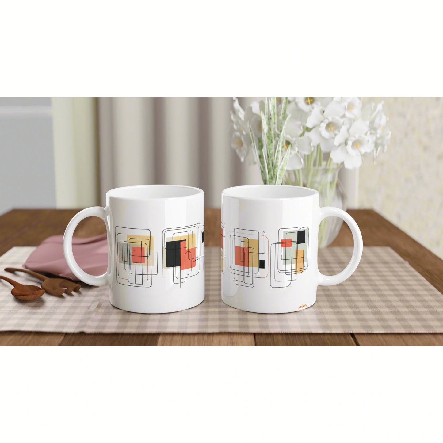 Two white mugs with geometric square-pattern designs on a checkered placemat; wooden table and a vase of white flowers in the background.