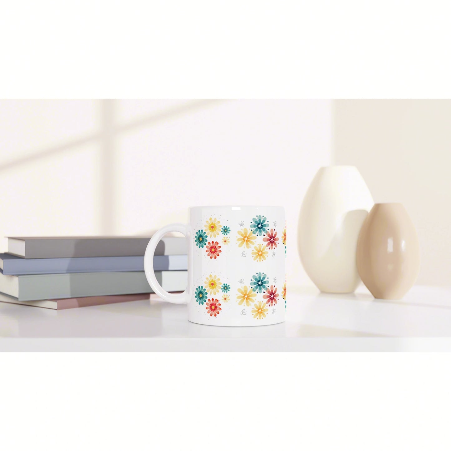 White mug with a bright multicolored flower pattern sits on a white surface beside stacked books and two ceramic vases.