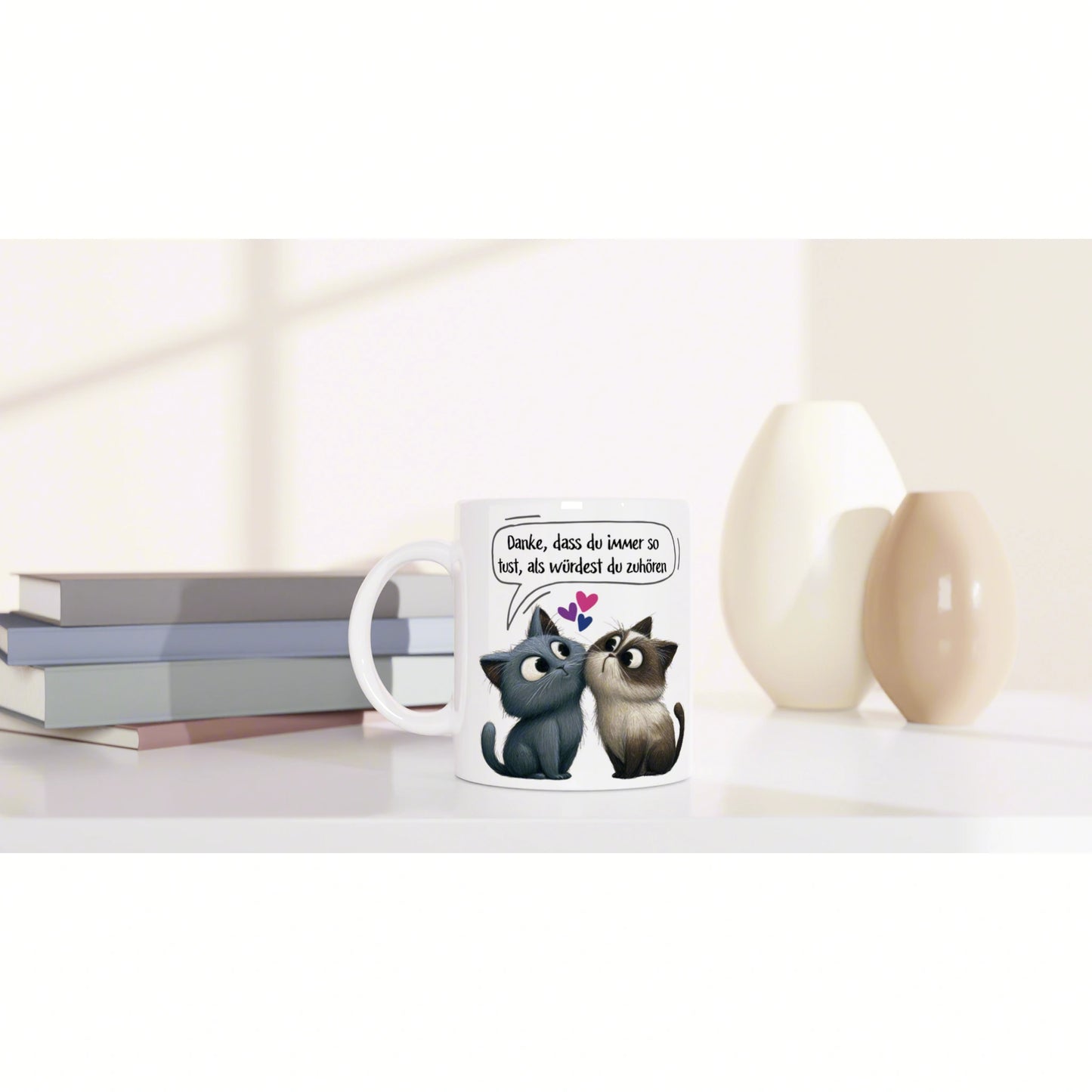 Product mockup: white mug with two cartoon cats, hearts, and a German speech bubble, on a desk beside books and decorative vases.