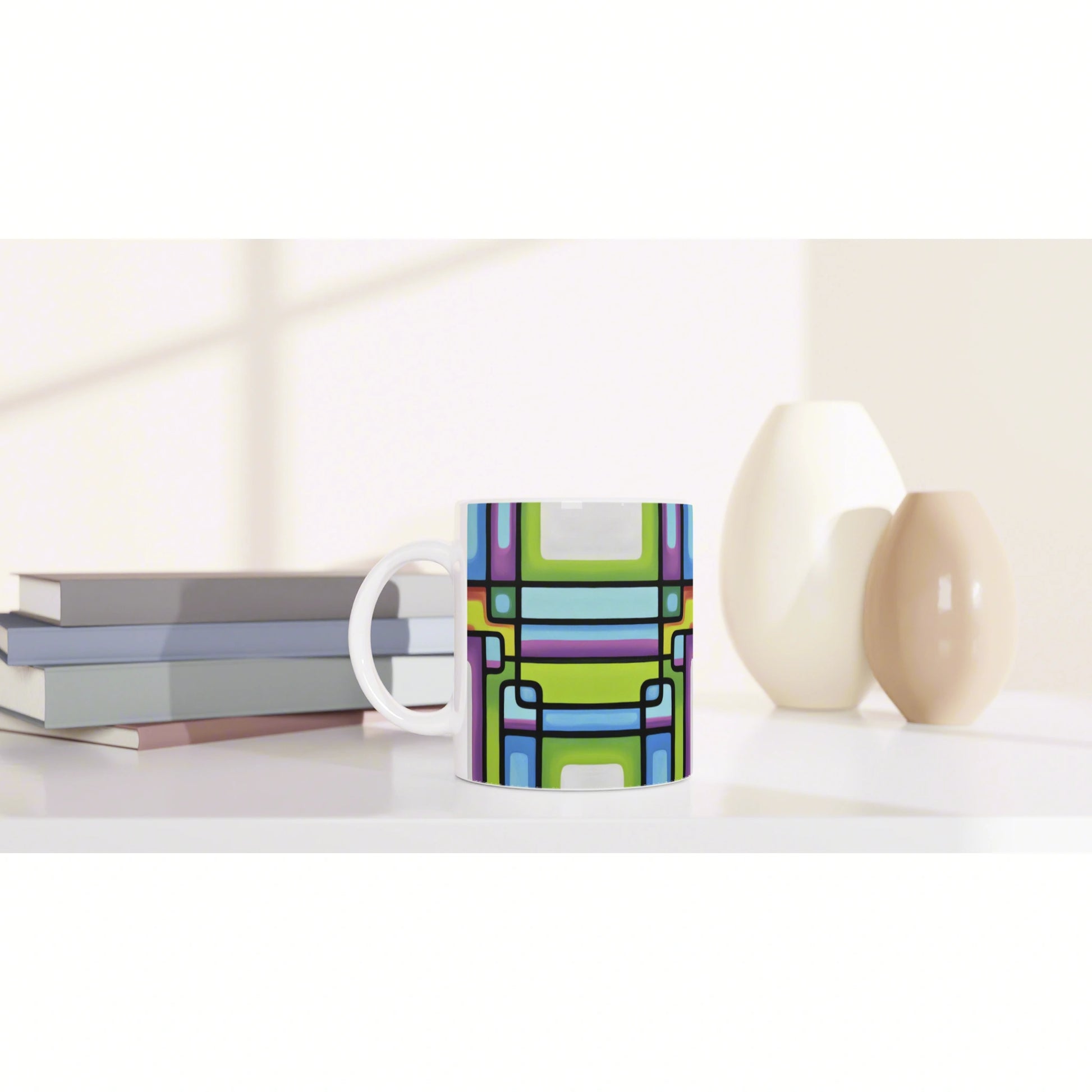 Product mockup: colorful geometric-pattern mug on a white desk with stacked books and ceramic vases.