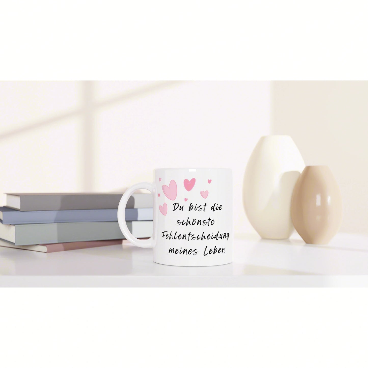White mug with pink hearts and a German slogan, 'Du bist die schönste Fehlentscheidung meines Lebens,' beside books and decorative vases on a pale desk.