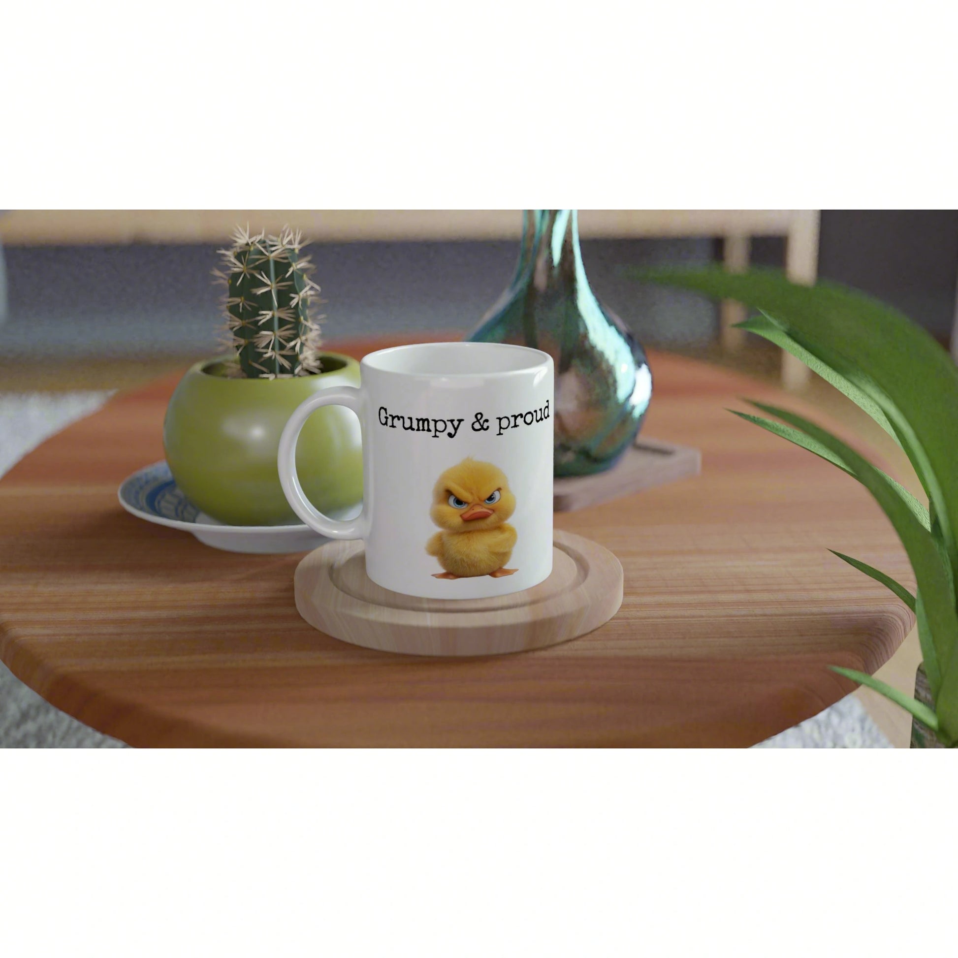 White ceramic mug with Grumpy & proud and a yellow chick on a wooden coaster, with plants in the background.
