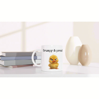 White mug with 'Grumpy & proud' and a grumpy yellow chick; sits on a desk beside books and decorative vases.