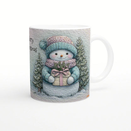 Illustrated mug with a smiling snowman in a teal knit hat and scarf, holding a wrapped gift among snowy evergreens and partial 'Merry Christmas' text.