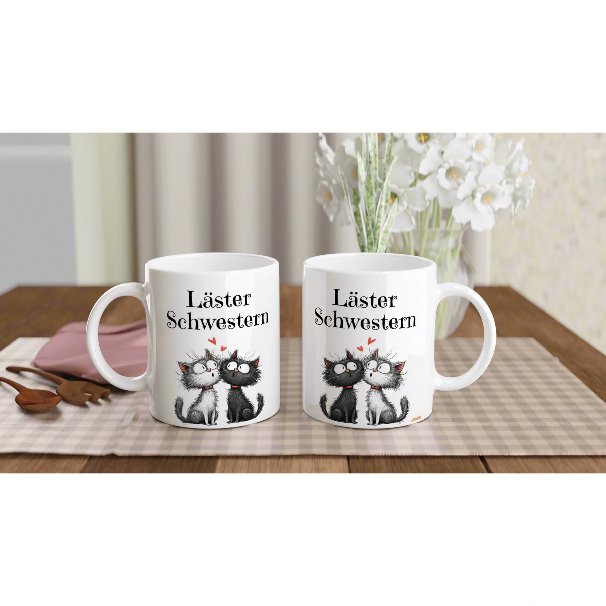 Two white mugs with cartoon cats and Läster Schwestern text on a plaid table runner with a vase of white flowers in the background.