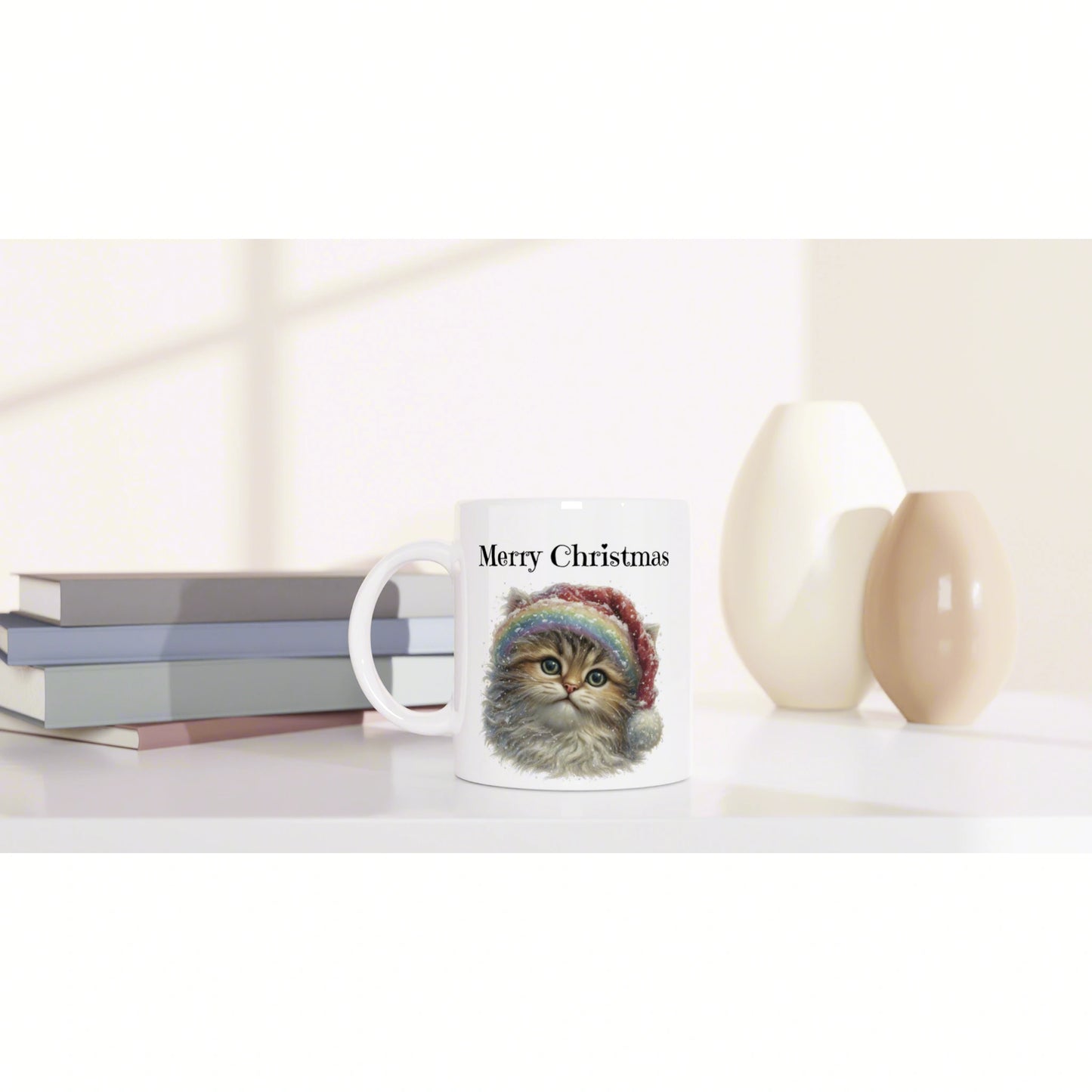 Product mockup: white mug with 'Merry Christmas' and a cute cat illustration, on a shelf with stacked books and decorative cream vases.
