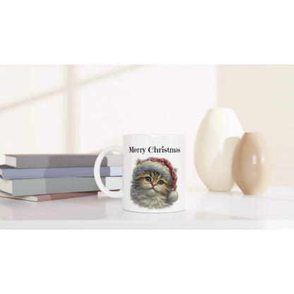 Product mockup: white mug with 'Merry Christmas' and a cute cat illustration, on a shelf with stacked books and decorative cream vases.