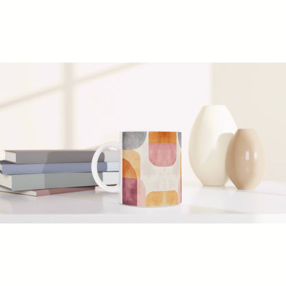 Product mockup: mug with a colorful geometric pattern on a white desk, beside stacked books and ceramic vases.