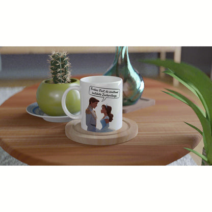 White mug with cartoon couple and a German speech bubble on a wooden coaster; cactus pot and teal vase on a wooden table.