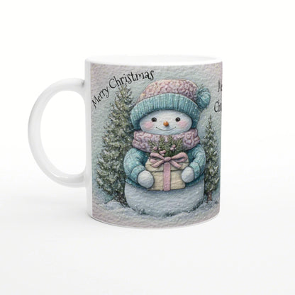 Mug with a cute snowman in teal hat and scarf holding a gift among snowy trees; Merry Christmas
