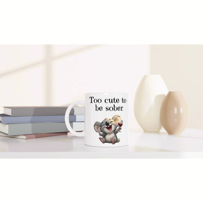 Mug mockup: white mug with 'Too cute to be sober' and a koala holding a wine glass, beside stacked books and vases.