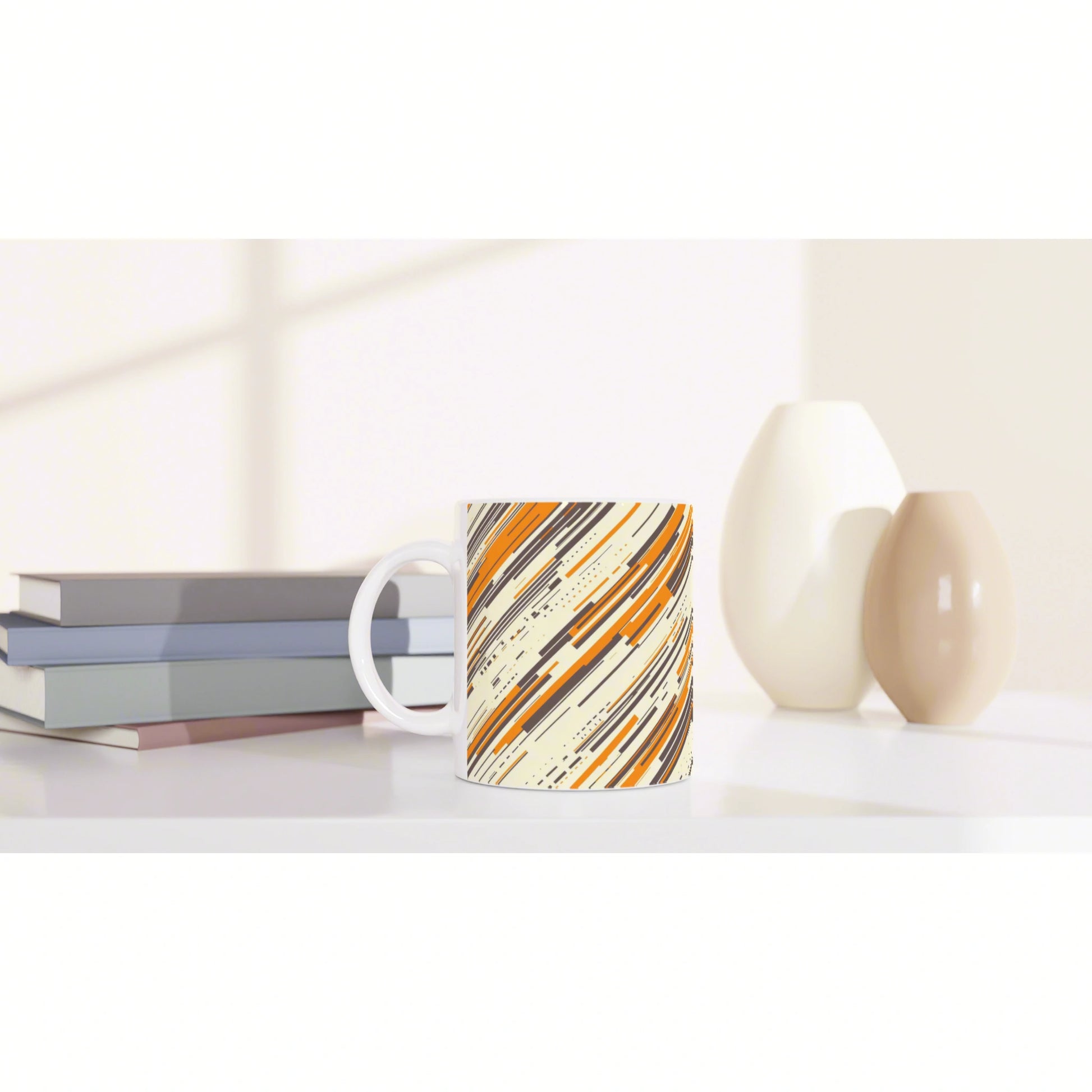 Mug with orange, cream, and black diagonal abstract pattern on a white desk, beside books and beige ceramic vases.