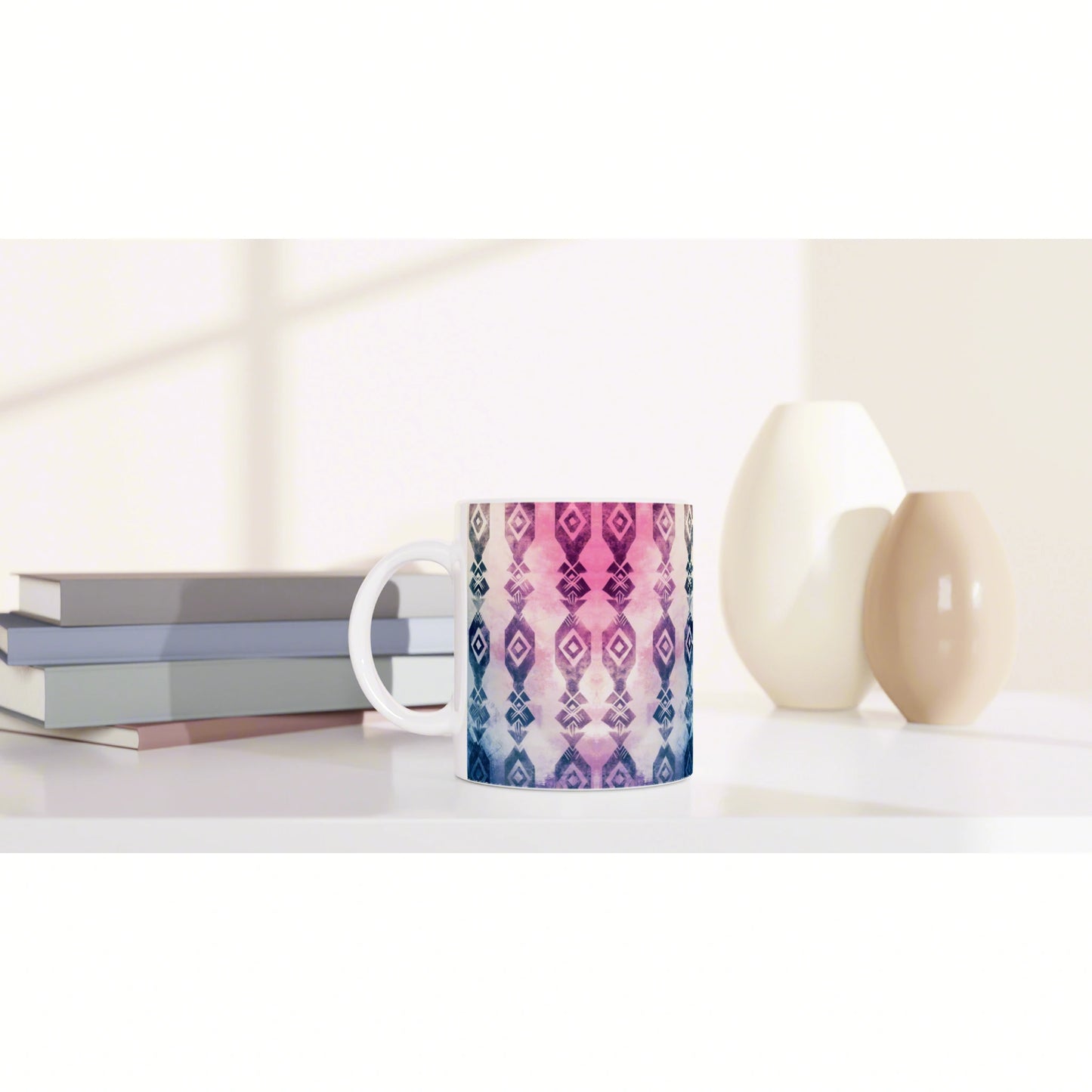 Mug featuring a pink-to-purple geometric pattern sits on a white desk beside pastel books and two ceramic vases.