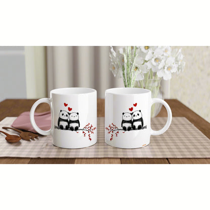 Two white mugs feature panda couple with red hearts on a checkered placemat, wooden table, with flowers in the background.