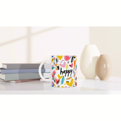 Product mockup: colorful mug with happy and brushstroke heart pattern, shown on a desk with books and ceramic vases.