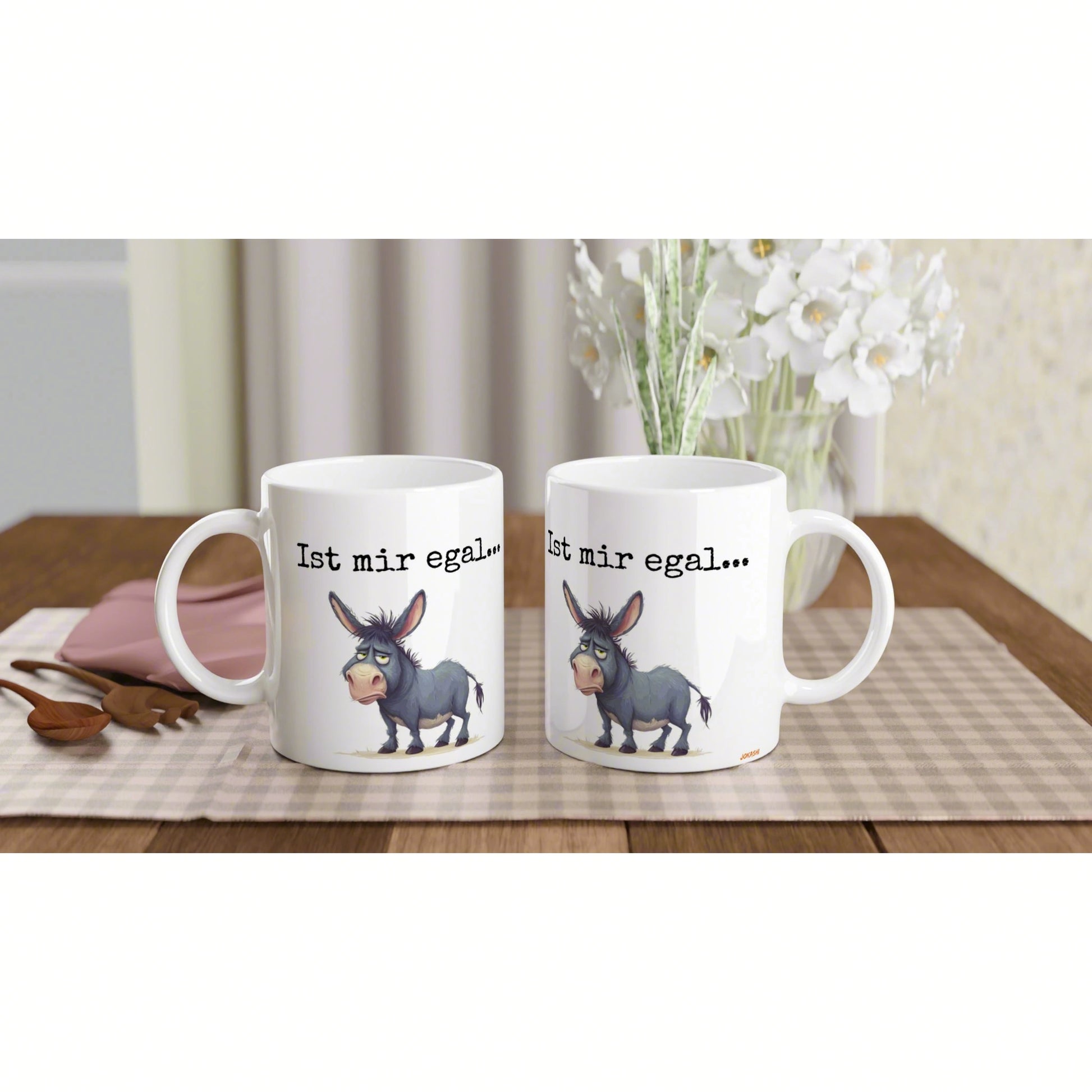 Product mockup: two white mugs with a cartoon donkey and the phrase 'Ist mir egal...' on a beige checkered table runner.