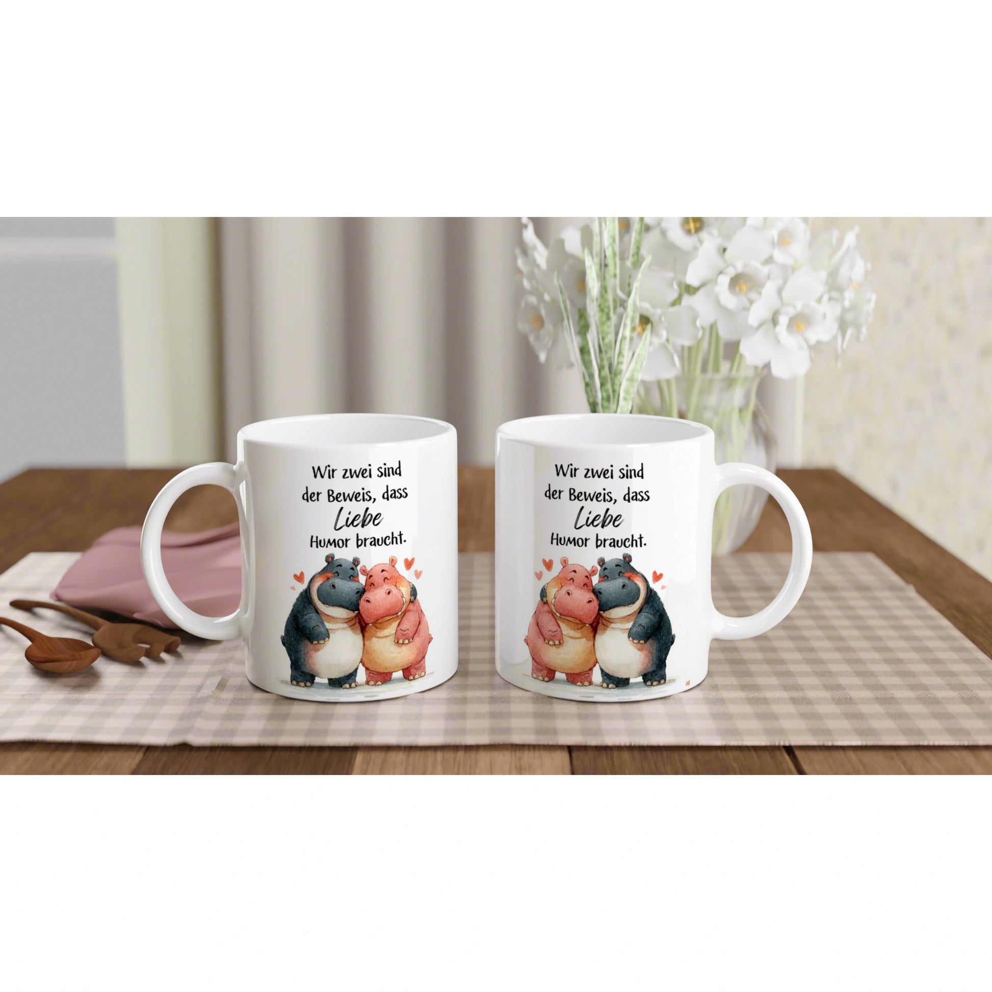Product mockup: two white mugs with cartoon hippos hugging and the German love-humor quote on a table.