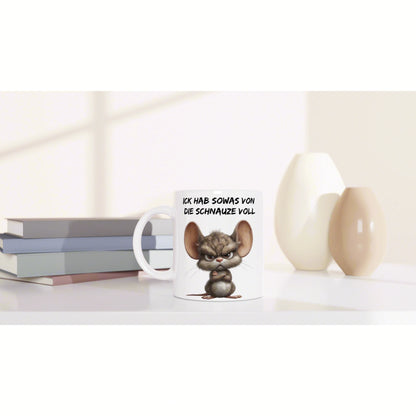 Product mockup: white mug with an angry cartoon mouse and German slogan, on a shelf with books and decorative vases.