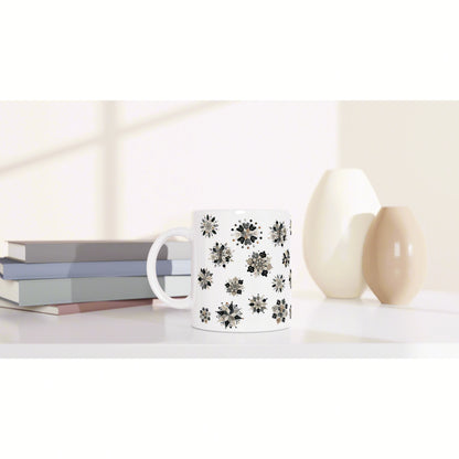 Product mockup: white mug with black floral pattern placed on a shelf beside pastel books and cream vases.
