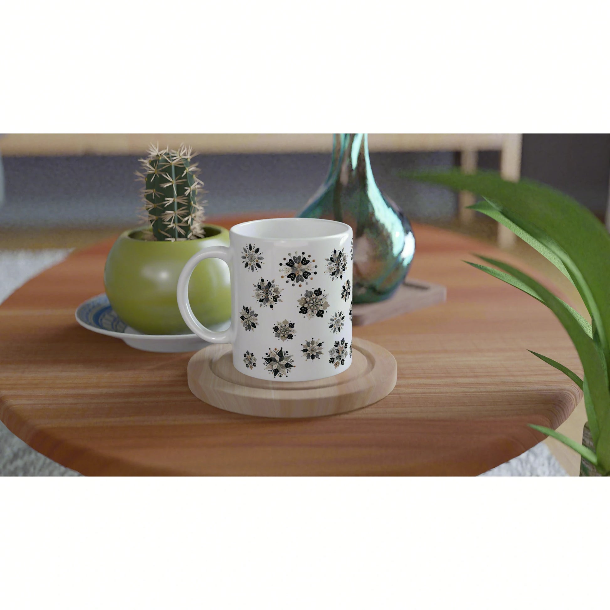 Product mockup: white mug with black floral pattern on a wooden table, beside a potted cactus and a decorative glass vase.