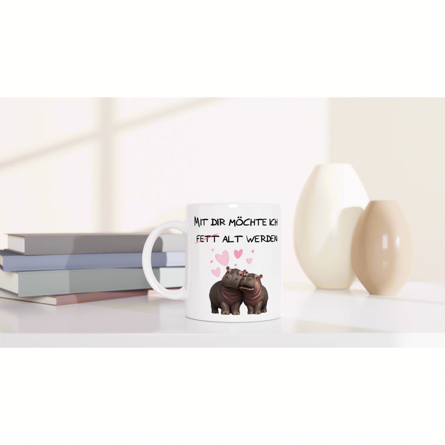 Product mockup: white mug with two hippos, pink hearts, and a German slogan, placed on a white desk near books and vases.