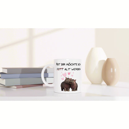 Product mockup: white mug with two hippos, pink hearts, and a German slogan, placed on a white desk near books and vases.