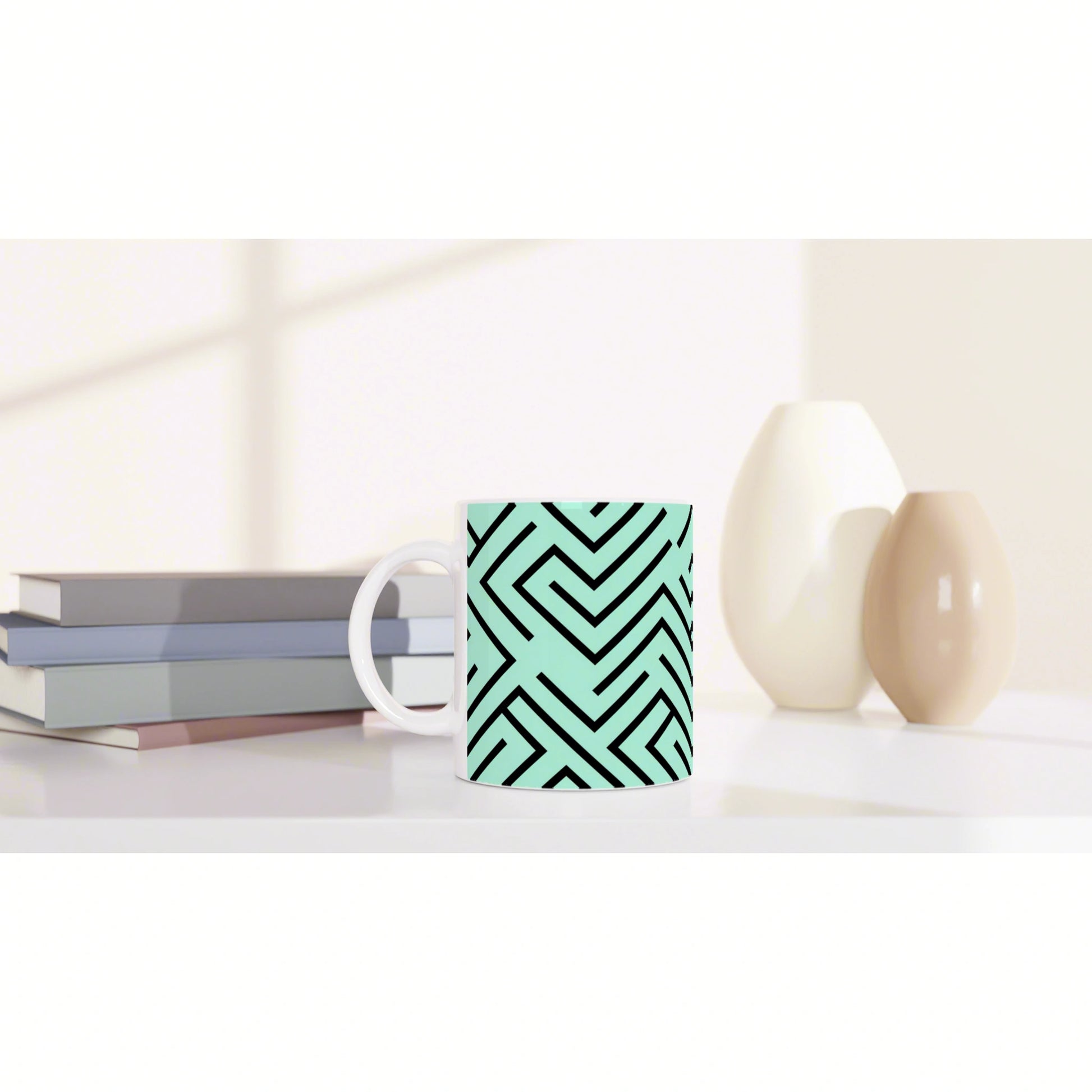 Mug with teal geometric maze pattern sits on a white shelf beside stacked books and white and beige vases.