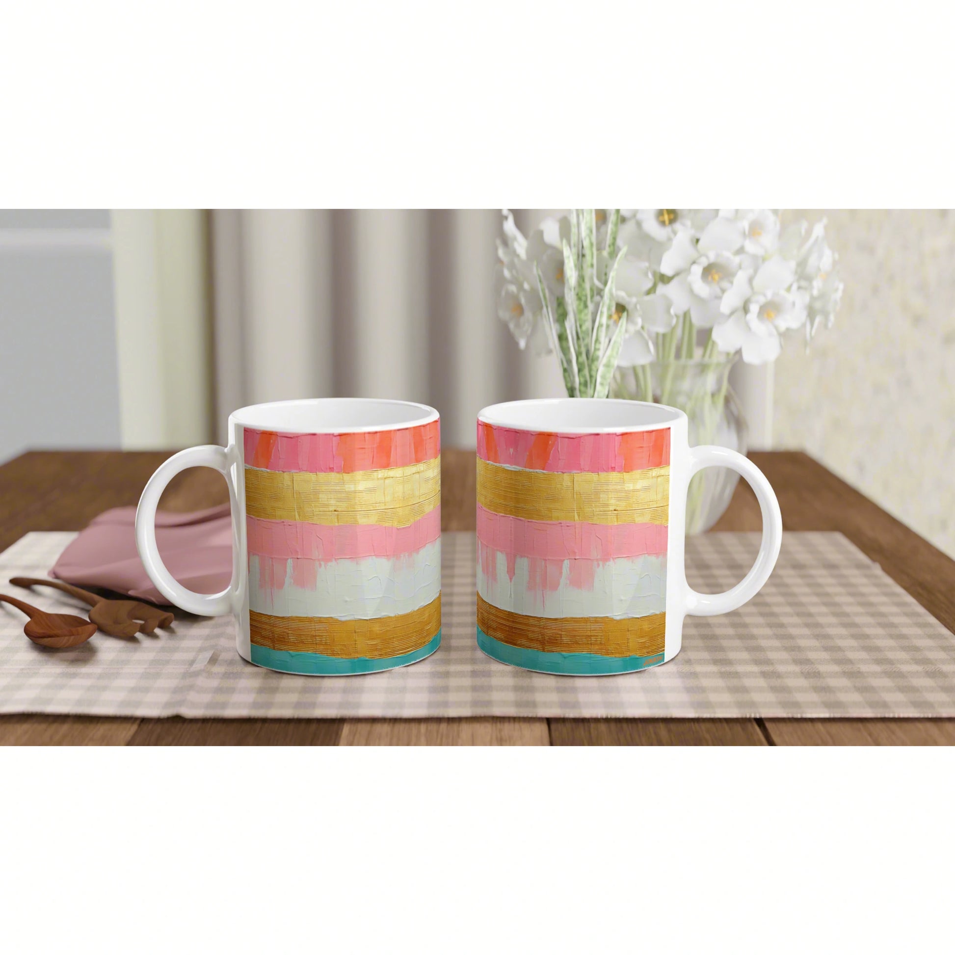 Two white ceramic mugs with colorful horizontal stripes (pink, gold, teal) on a checkered placemat; blurred vase of white flowers in the background.