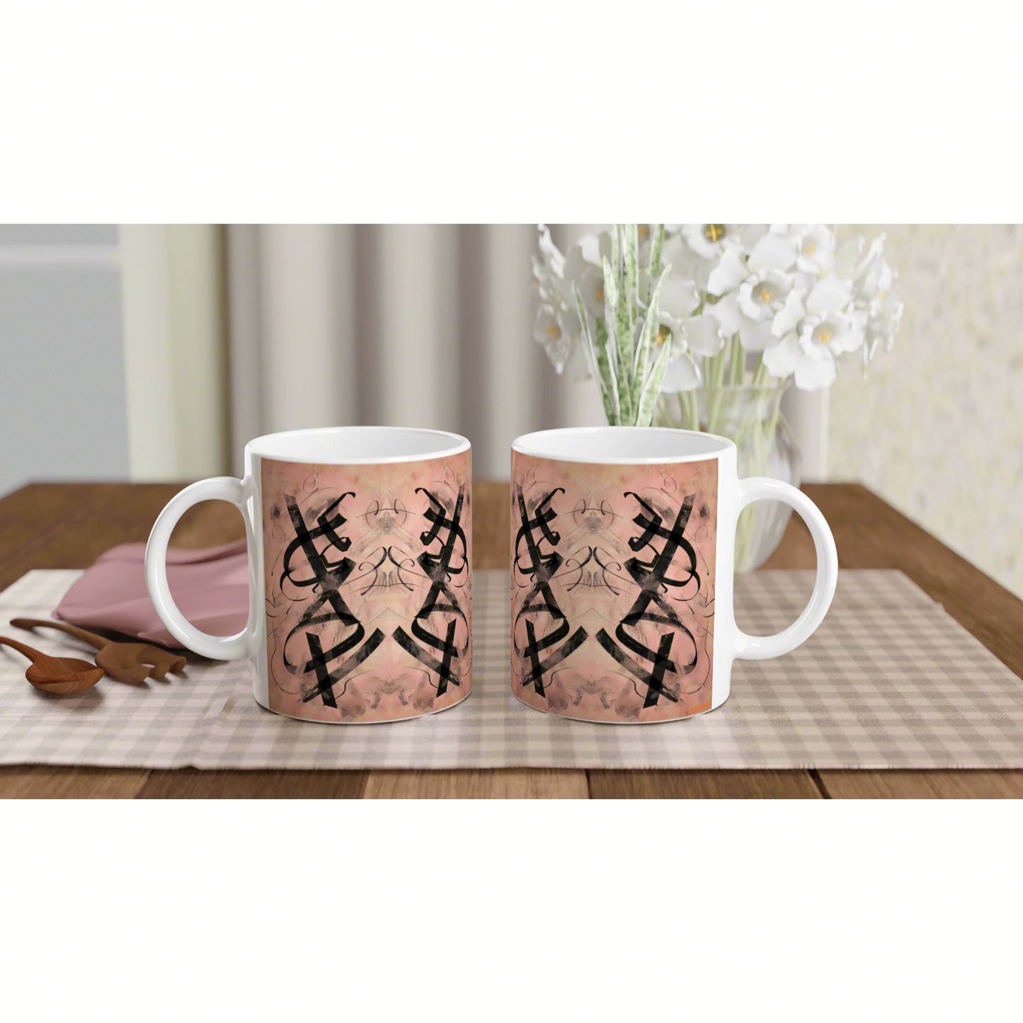 Two white ceramic mugs on a wooden table featuring a pink patterned wrap with black mirrored geometric motifs.
