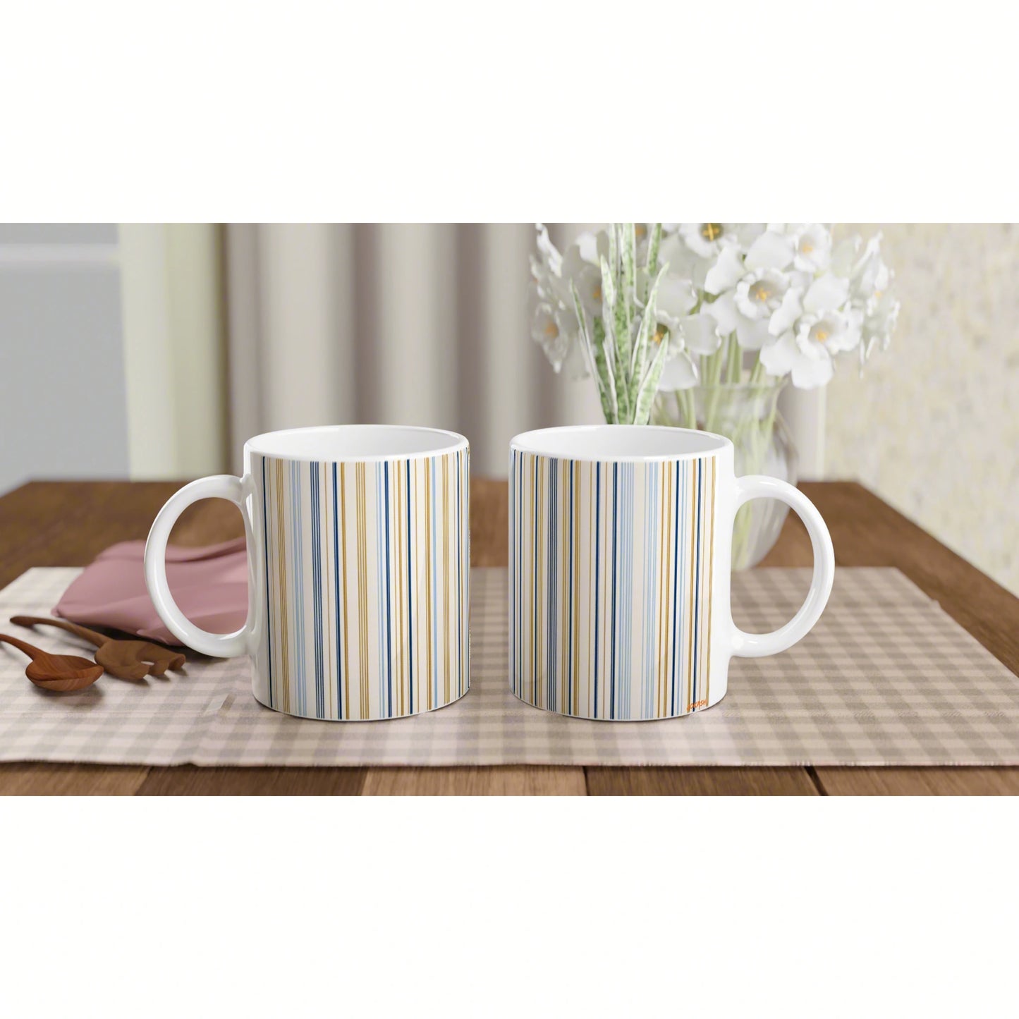 Two white ceramic mugs with vertical blue, gold, and beige stripes on a wooden table; white flowers in a vase in the background.