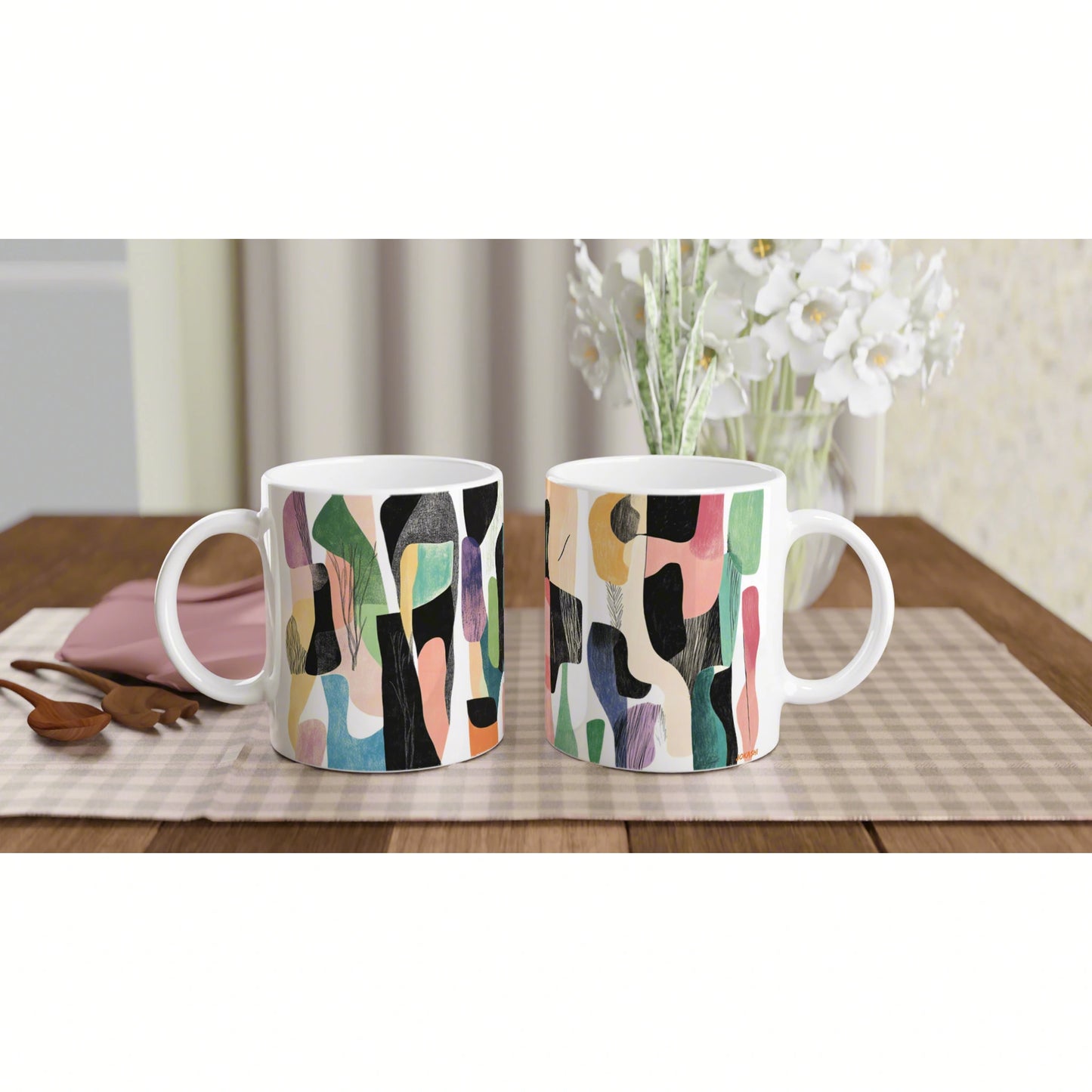 Two white ceramic mugs with abstract multicolor patterns on a beige placemat on a wooden table.