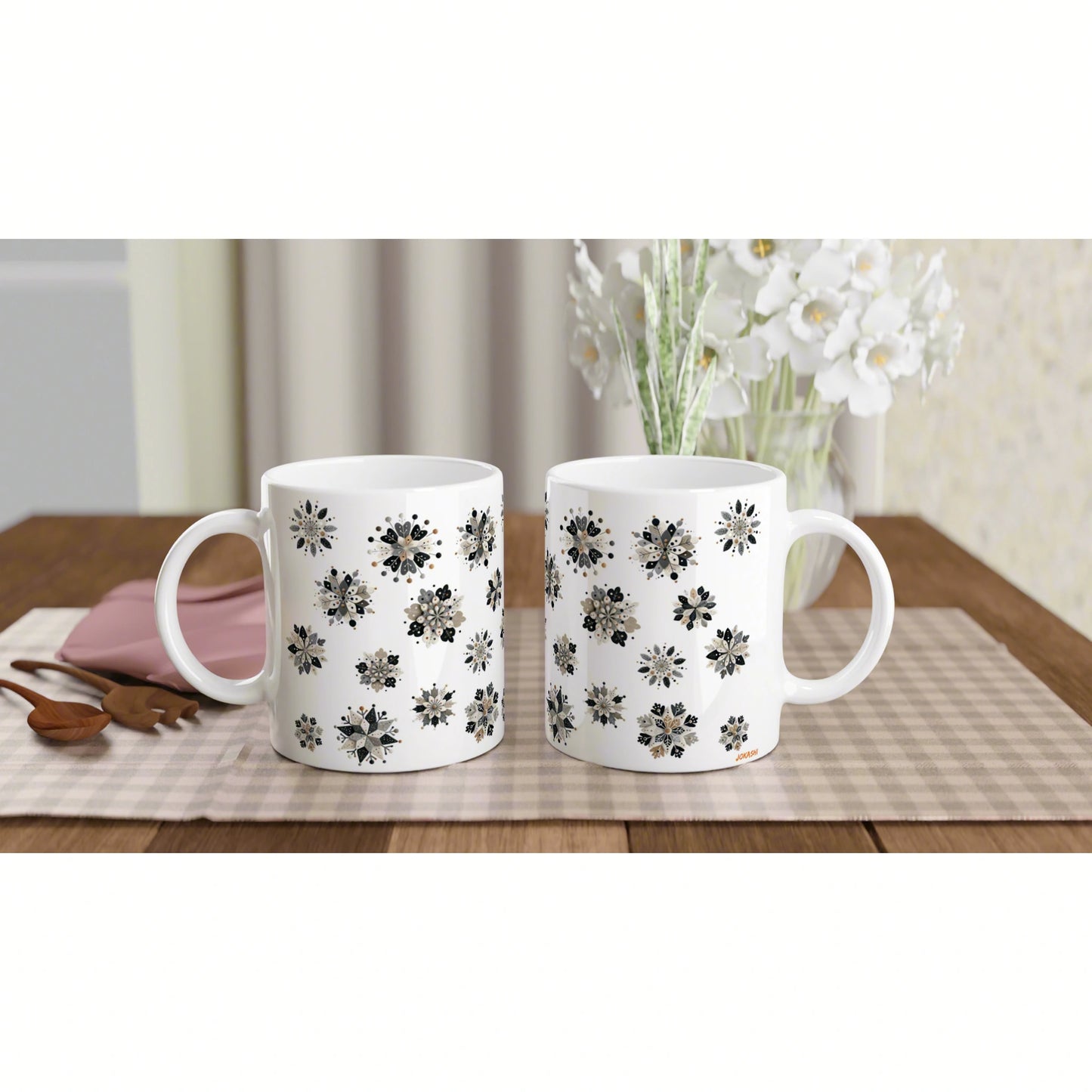 Two white ceramic mugs with black floral patterns sit on a beige checkered placemat on a wooden table beside white flowers.