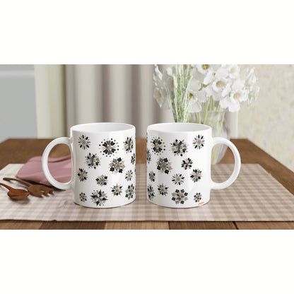 Two white ceramic mugs with black floral patterns sit on a beige checkered placemat on a wooden table beside white flowers.