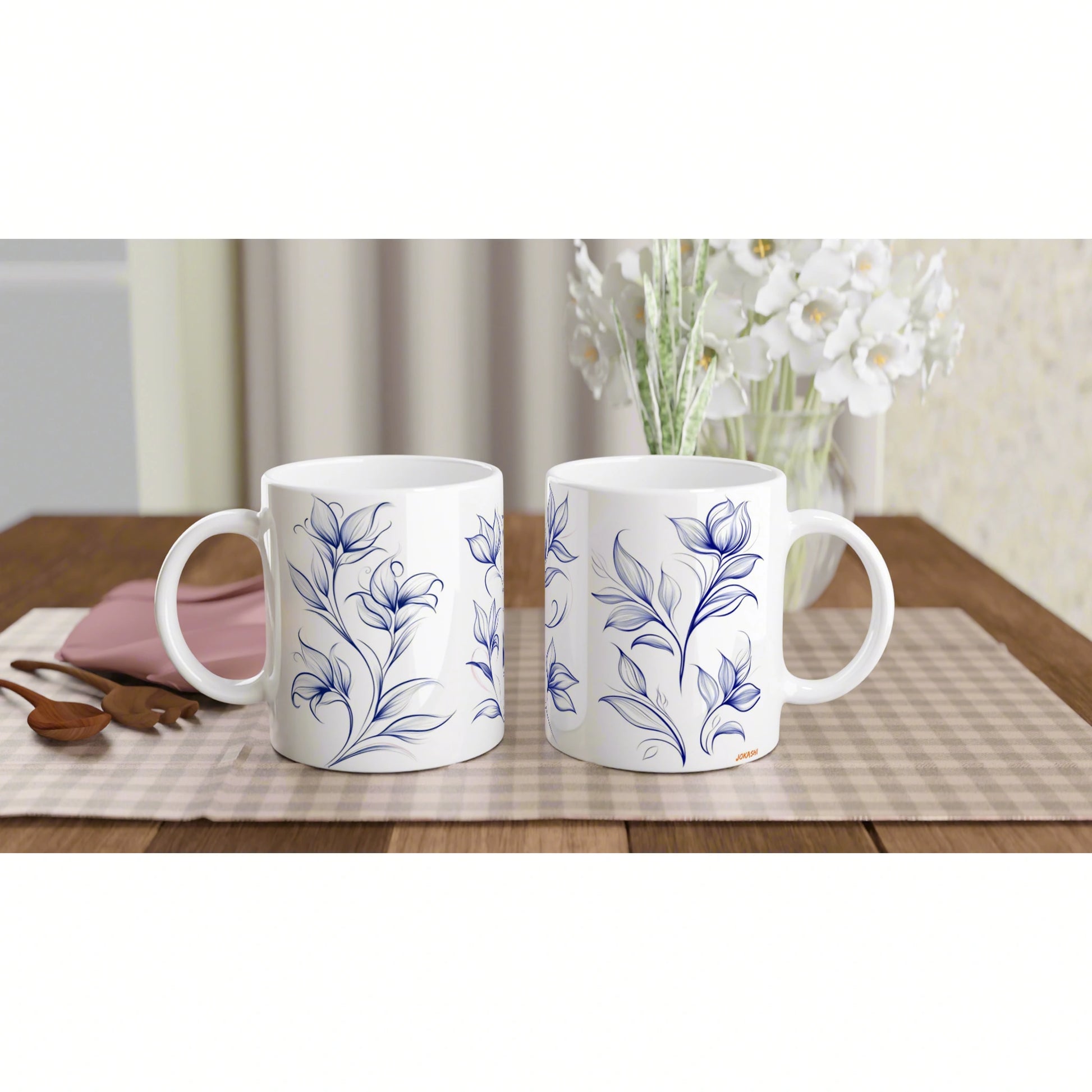 Two white ceramic mugs with blue floral patterns on a wooden table, with a vase of white flowers in the blurred background.