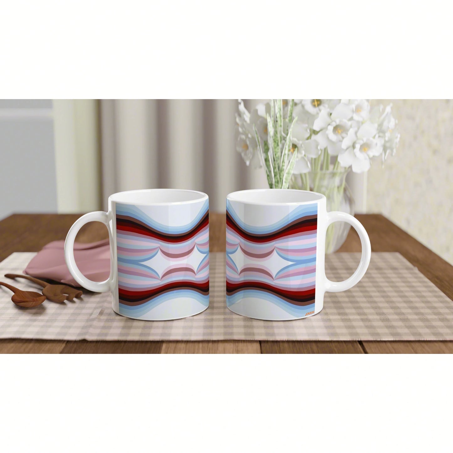 Two white ceramic mugs with a blue-red abstract wave pattern on a wooden table; beige placemat and background flowers.