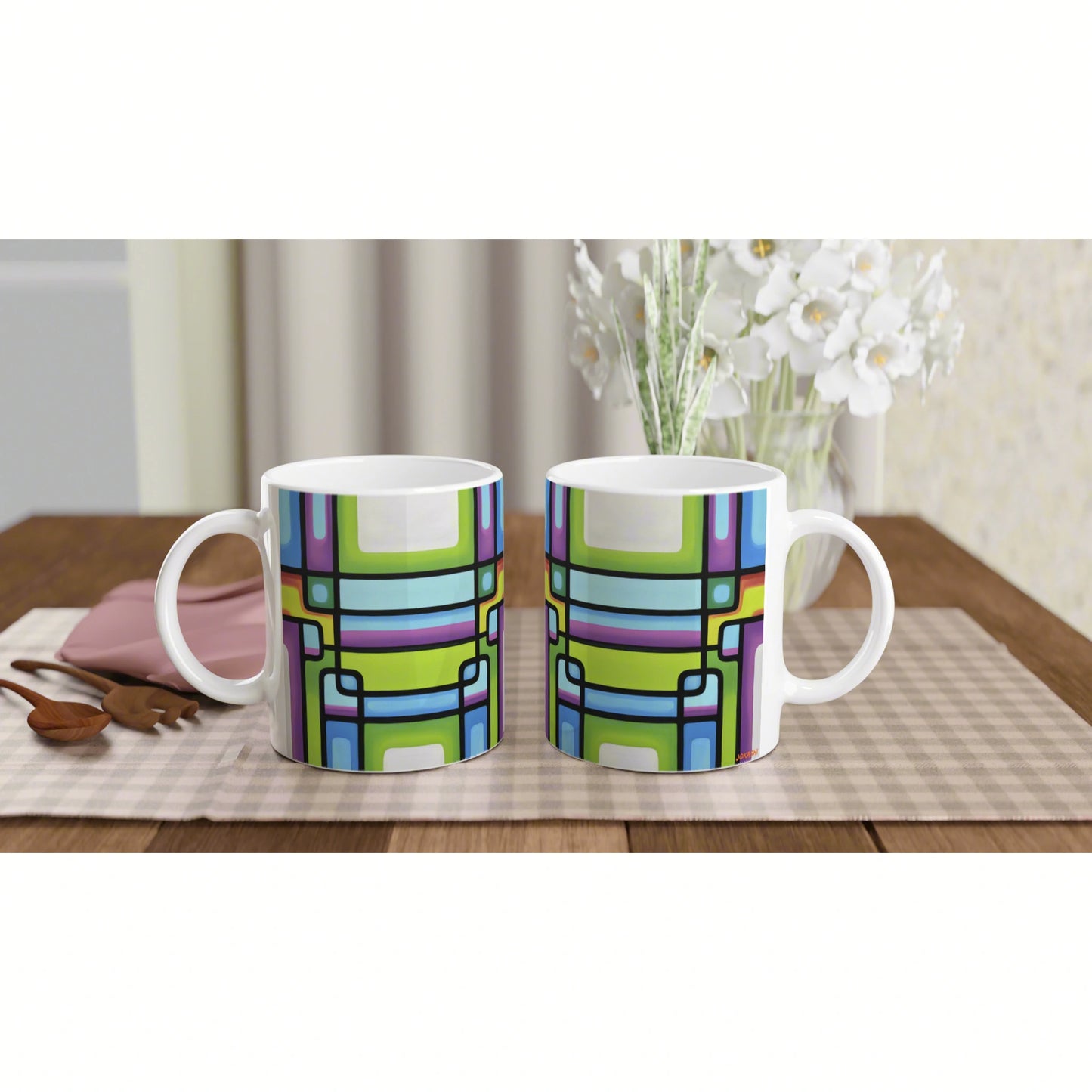 Two white ceramic mugs with a bright geometric multicolor pattern on a wooden table, placed on a plaid placemat with white flowers in the background.