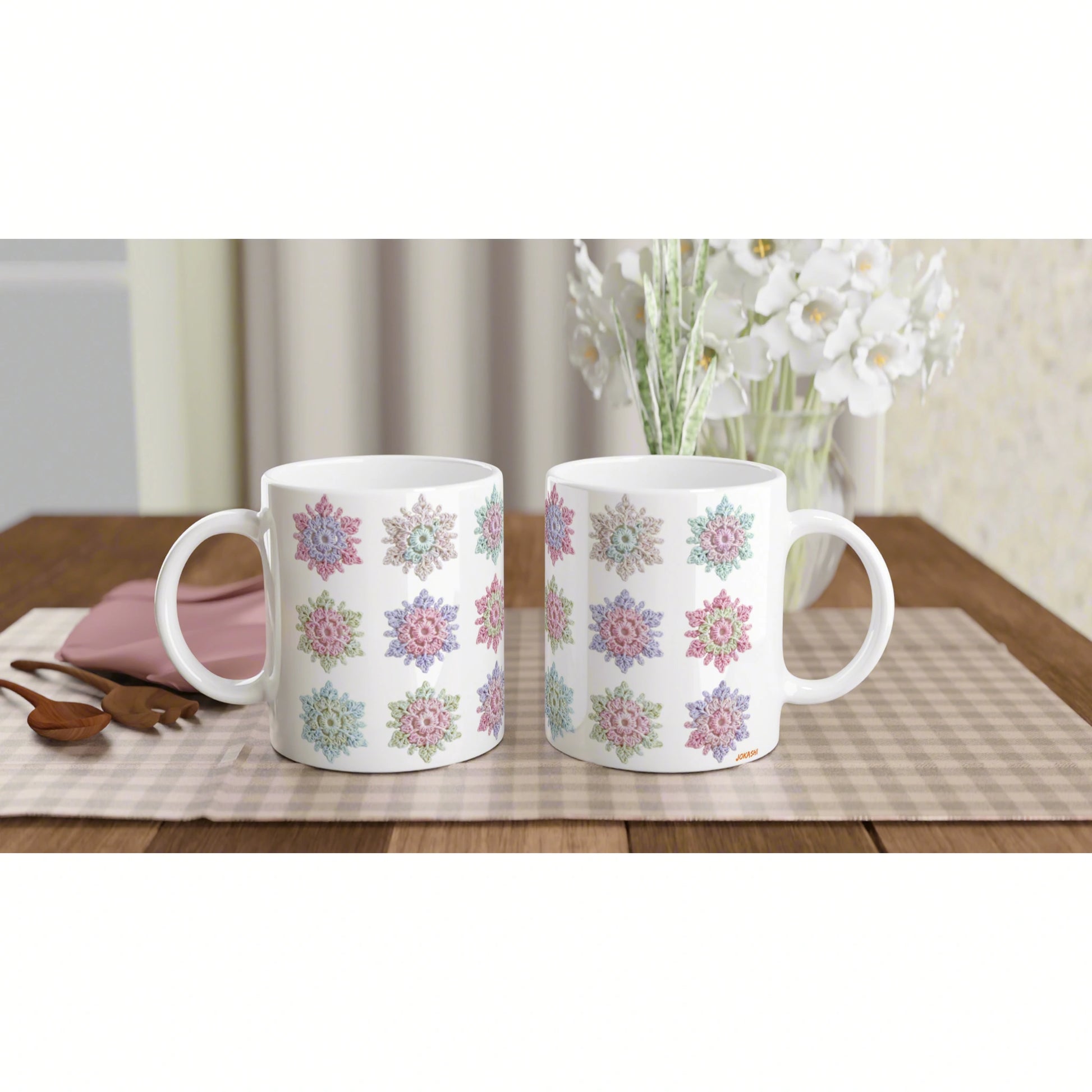 Two white ceramic mugs with colorful floral patterns on a wooden table; a vase of white flowers in the blurred background.