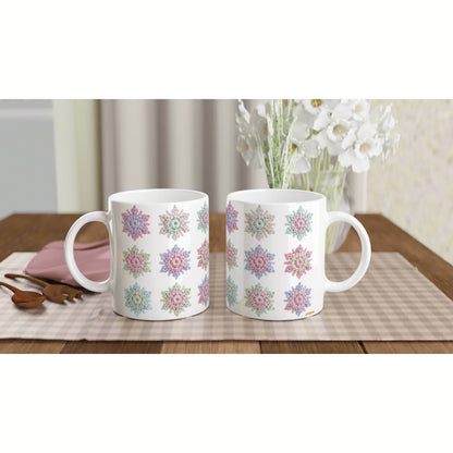 Two white ceramic mugs with colorful floral patterns on a wooden table; a vase of white flowers in the blurred background.