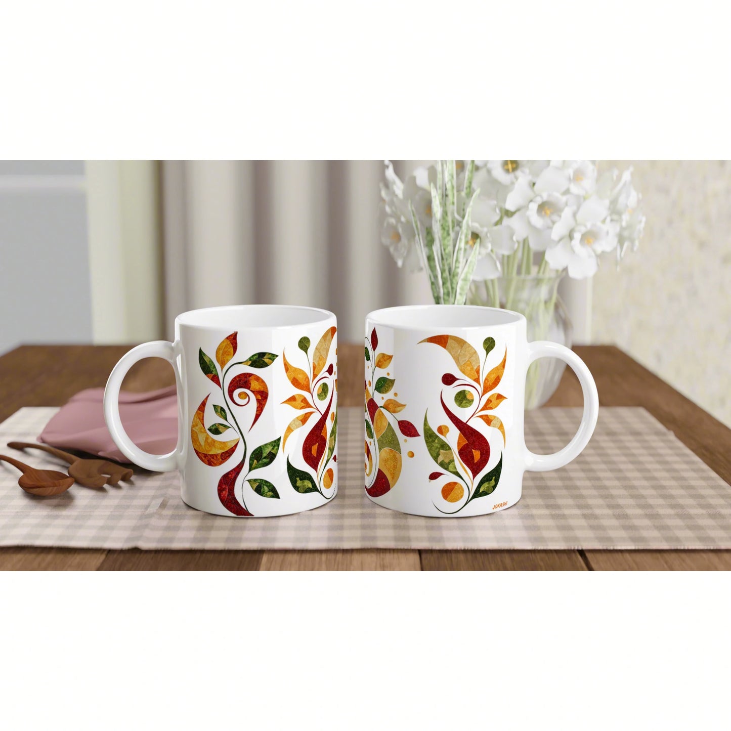 Two white ceramic mugs with colorful leafy patterns on a checkered mat; wooden table, white floral vase in background.