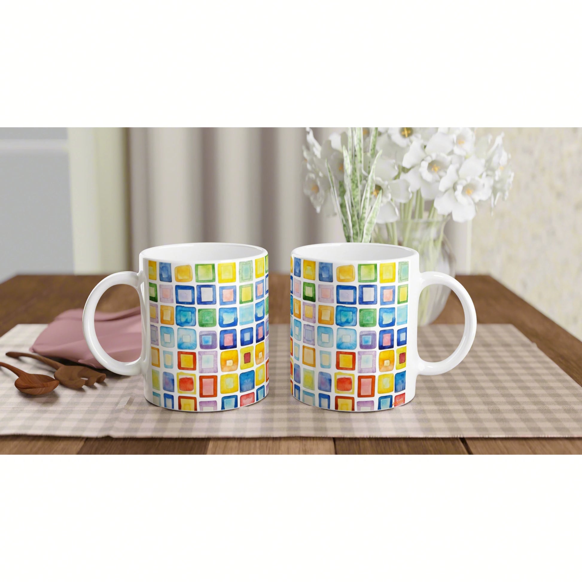 Two white ceramic mugs with a colorful square mosaic pattern on a wooden table, with a vase of white flowers in the background.
