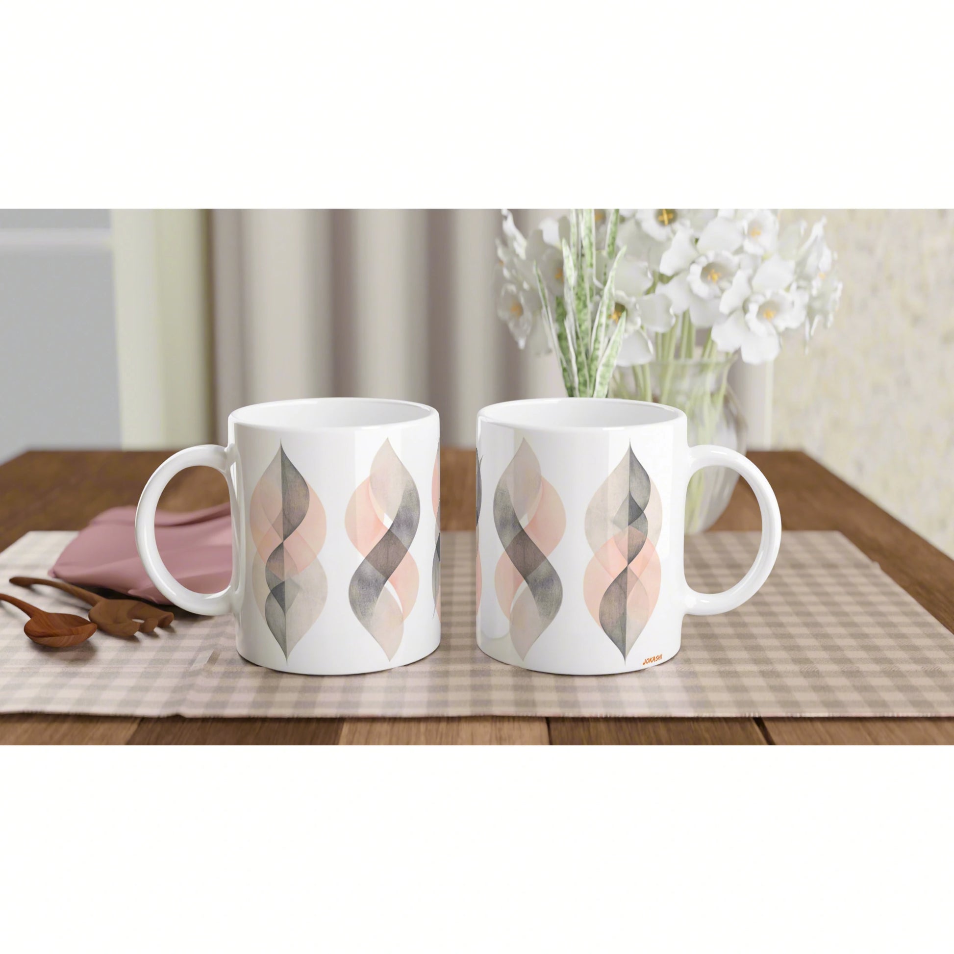 Two white ceramic mugs with a pastel geometric leaf pattern in pink, gray, and beige on a checkered placemat, with white flowers.