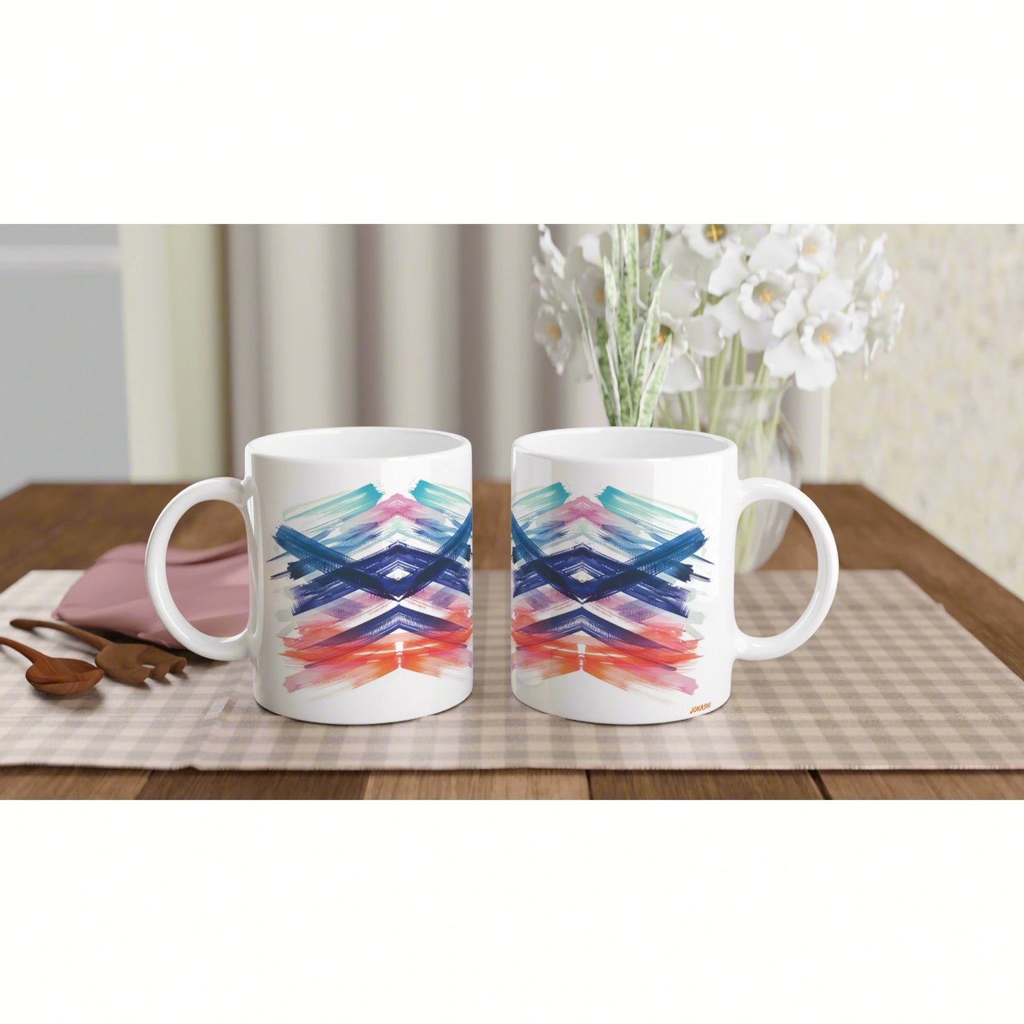 Two white mugs with blue, teal, pink and orange abstract brushstrokes on a wooden table; white flowers in the background.