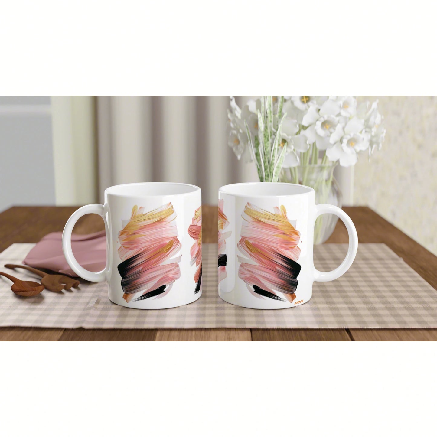 Two white mugs with pink, orange, and black brushstroke patterns sit on a checkered placemat on a wooden table.