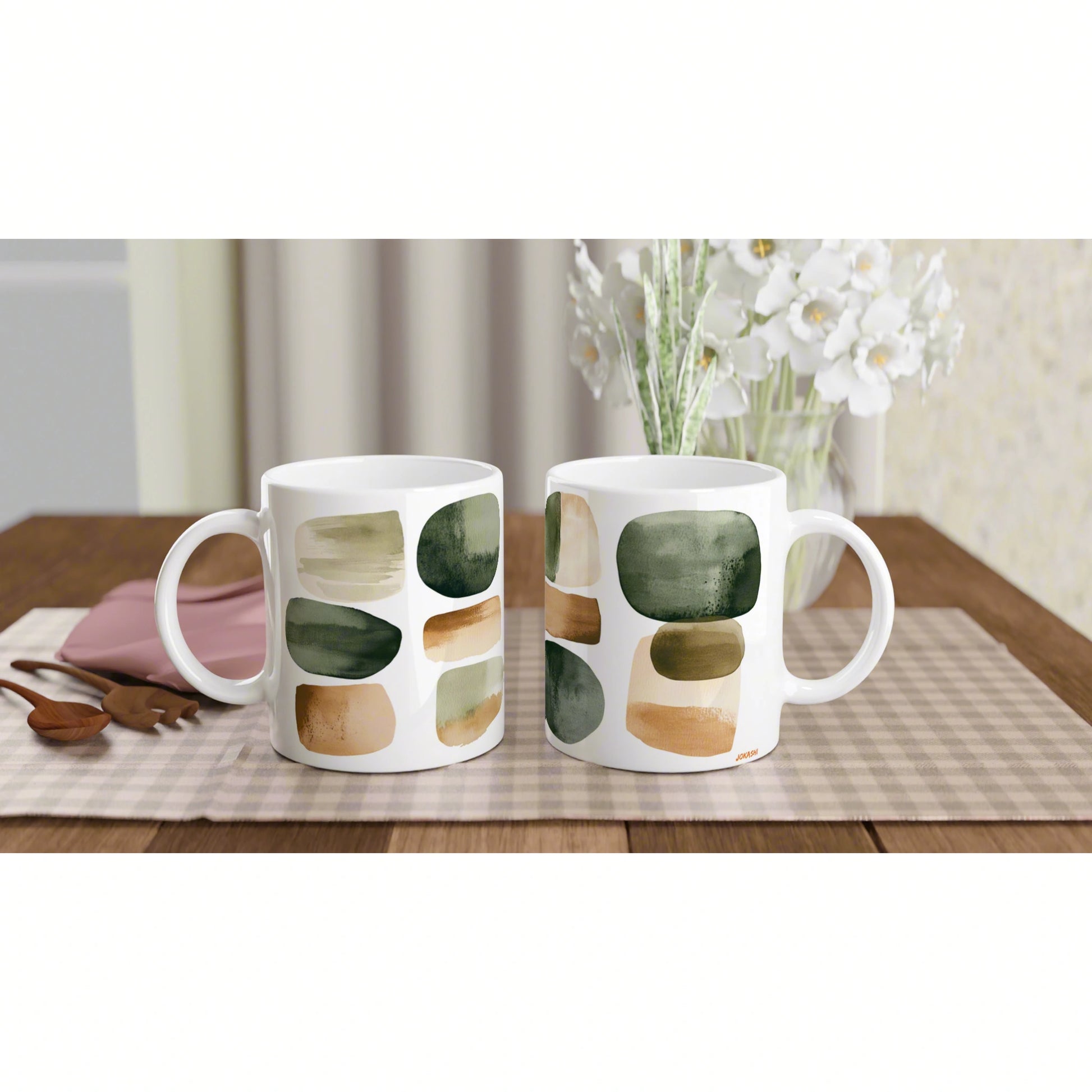 Two white mugs with abstract green and beige brushstrokes on a wooden table, with a vase of white flowers in the background.