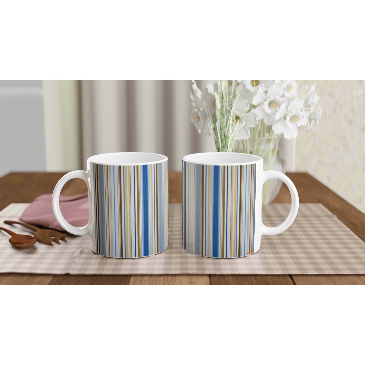 Two white mugs with blue and beige vertical stripes sit on a wooden table with a vase of white flowers in the background.