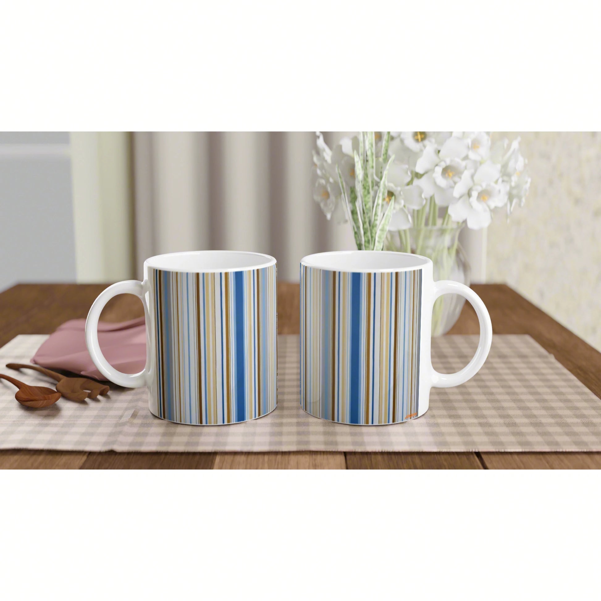 Two white mugs with blue and beige vertical stripes sit on a wooden table with a vase of white flowers in the background.