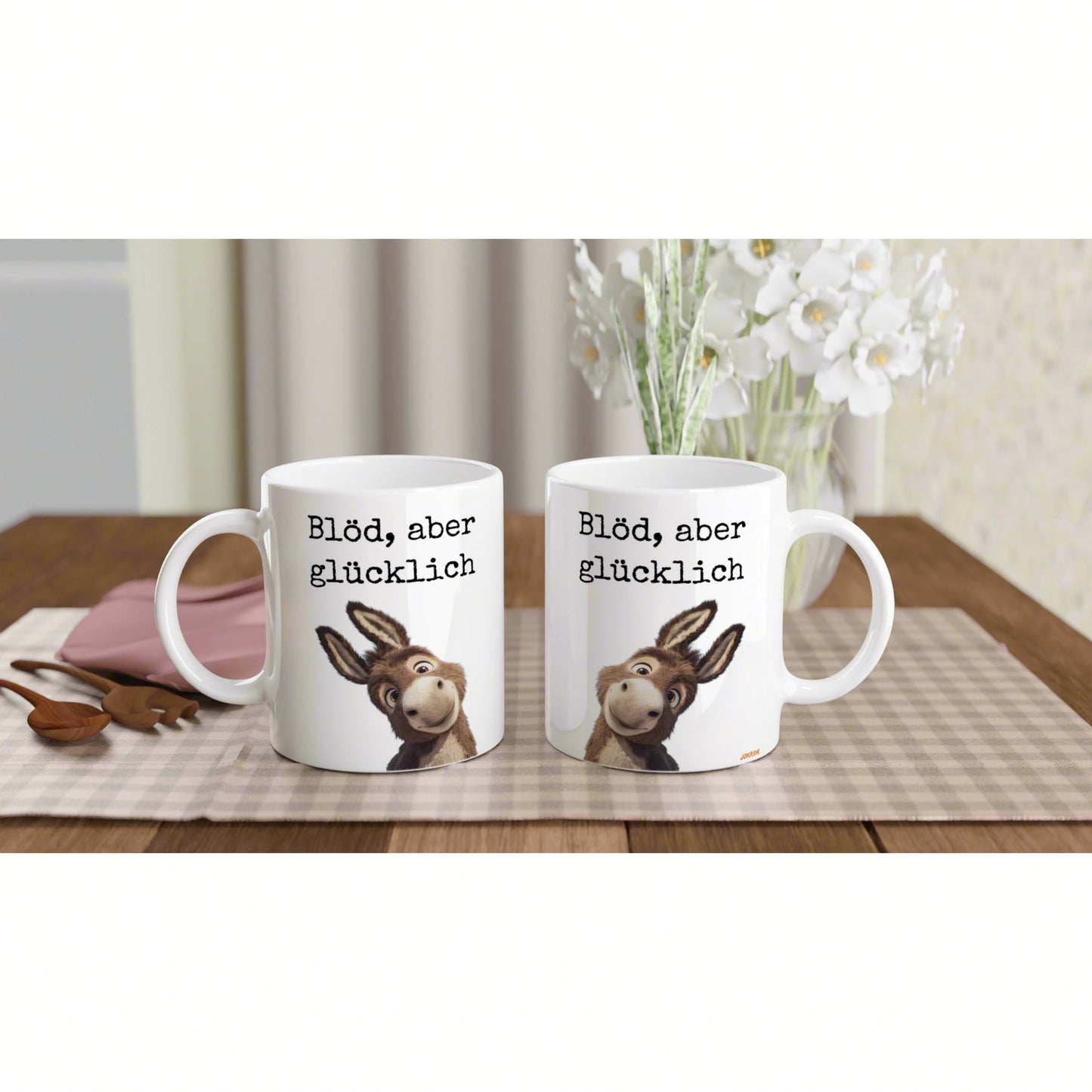 Two white mugs with a cartoon donkey and the German phrase 'Blöd, aber glücklich' on a beige checkered placemat.